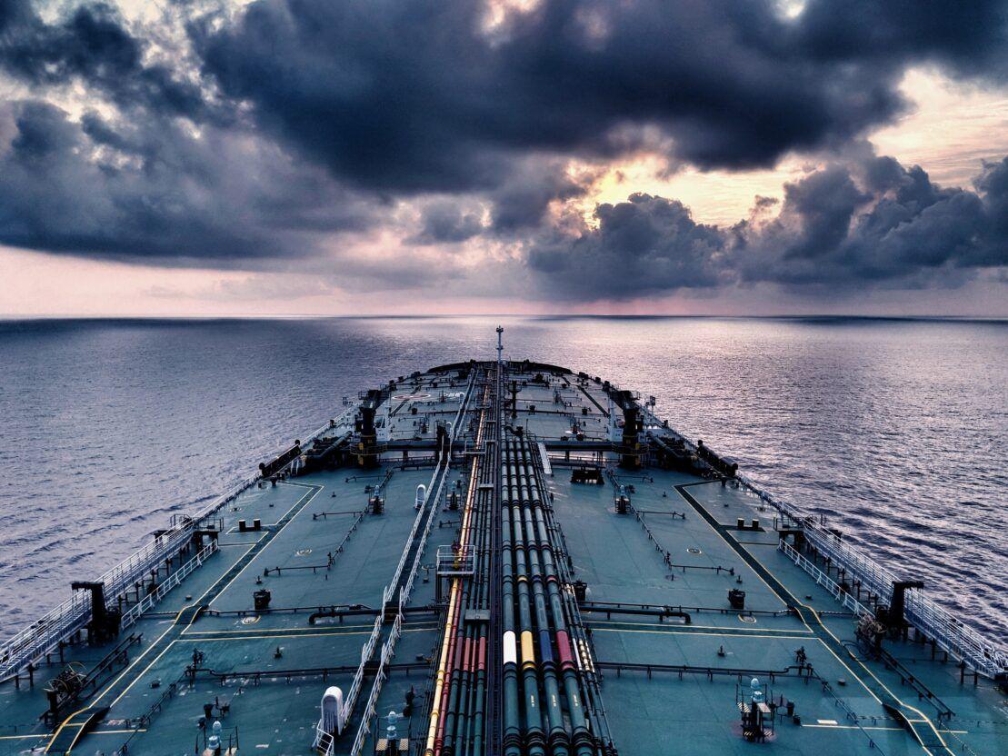 View from the stern along the deck of an oil tanker at sea, with cargo pipelines in the foreground and a cloudy sunset sky.