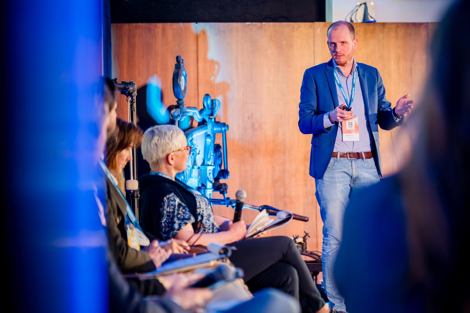 Un ponent intervé dret a l'escenari del BlueInvest Day 2026 davant d'un panell d'expertes assegudes durant una sessió de l'esdeveniment a Brussel·les.