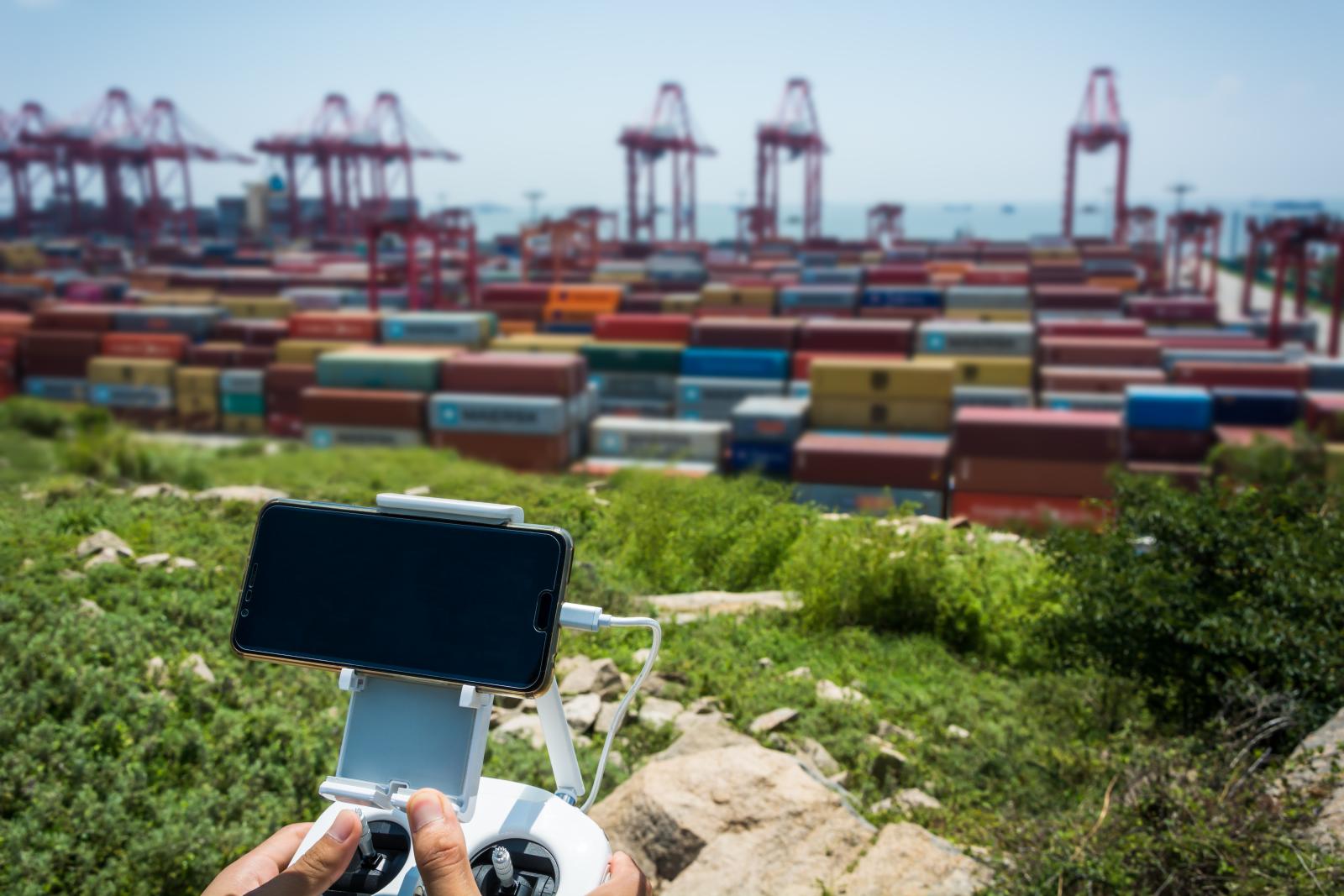 Persona maneja el mando de un dron, con un smartphone acoplado como pantalla, frente a una terminal de contenedores apilados y grúas pórtico rojas al fondo.