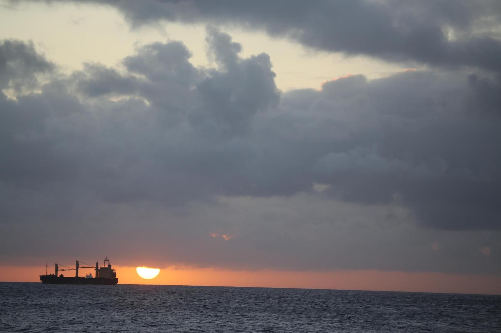 Silueta de un buque de carga navegando en el horizonte durante la puesta de sol, con un cielo nublado
