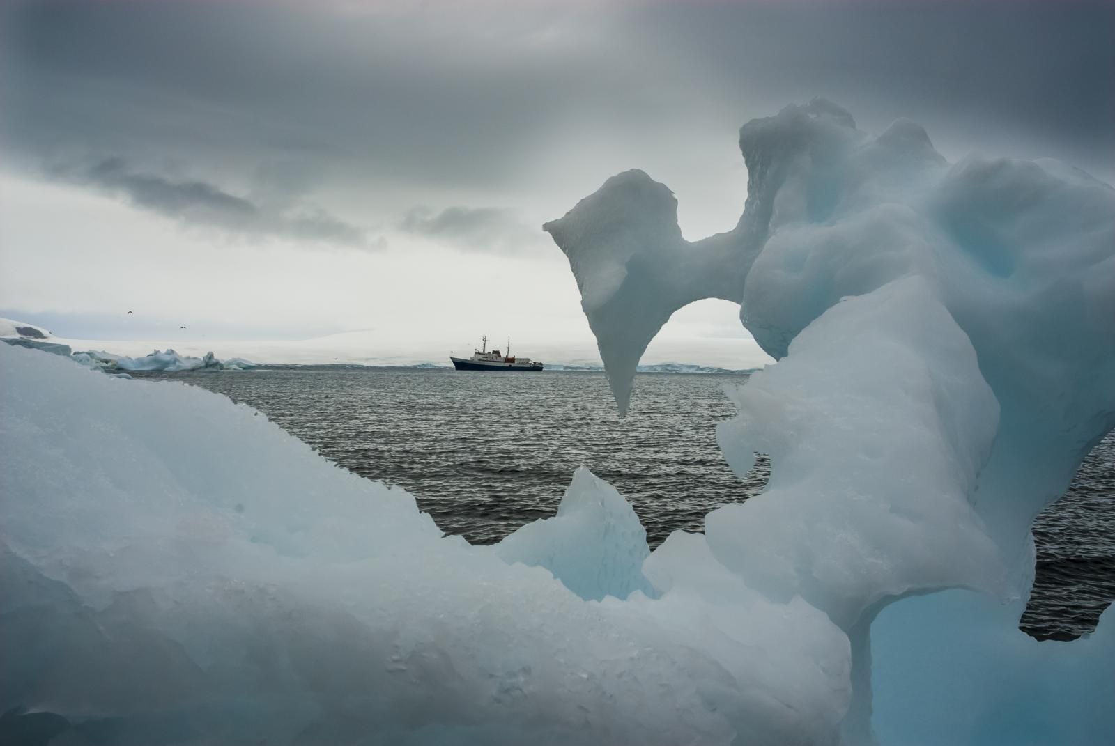 A ship sails through polar waters, seen through a gap between blocks of ice, under an overcast sky.