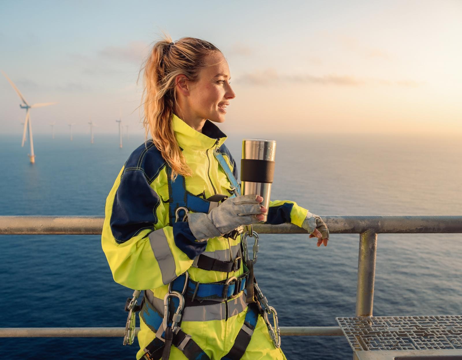 Técnica con mono reflectante y arnés de seguridad sostiene un termo en una plataforma marina, con aerogeneradores al fondo.