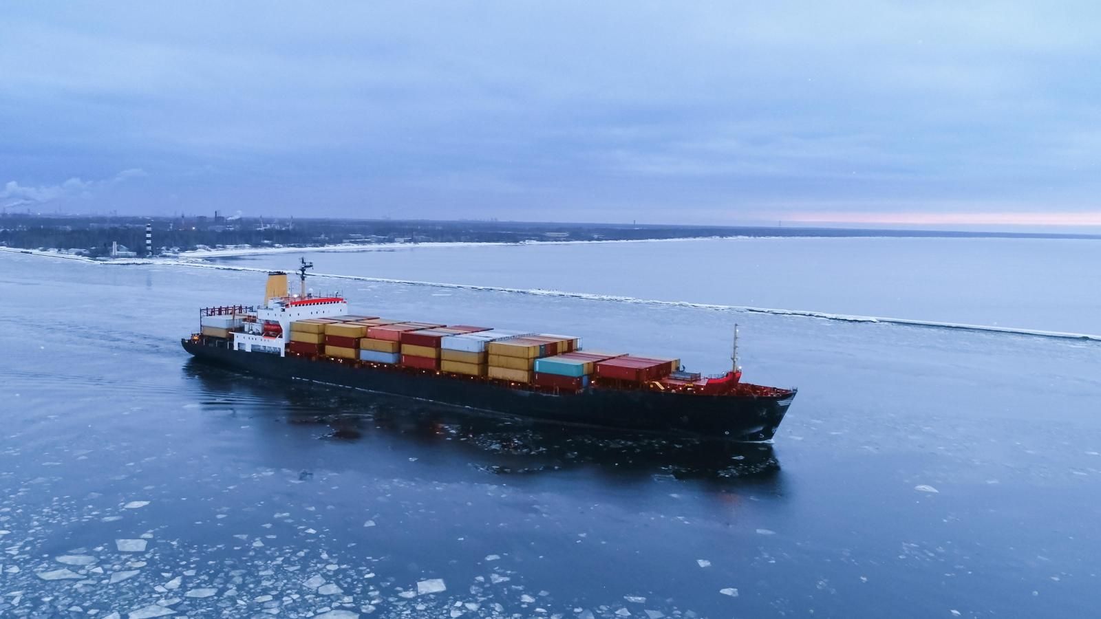 Aerial view of a red-hulled icebreaker on an expanse of sea ice, with the channel it has cut curving away behind it toward open water and snow-covered mountains on the horizon.