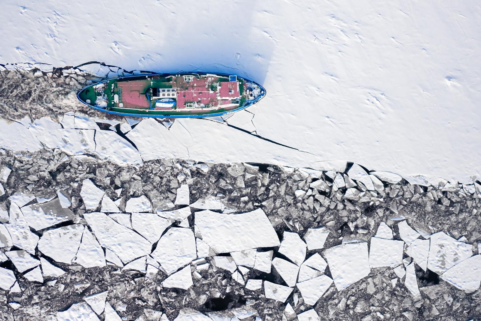Overhead view of an icebreaker advancing through an ice sheet that fractures into floes in its wake.
