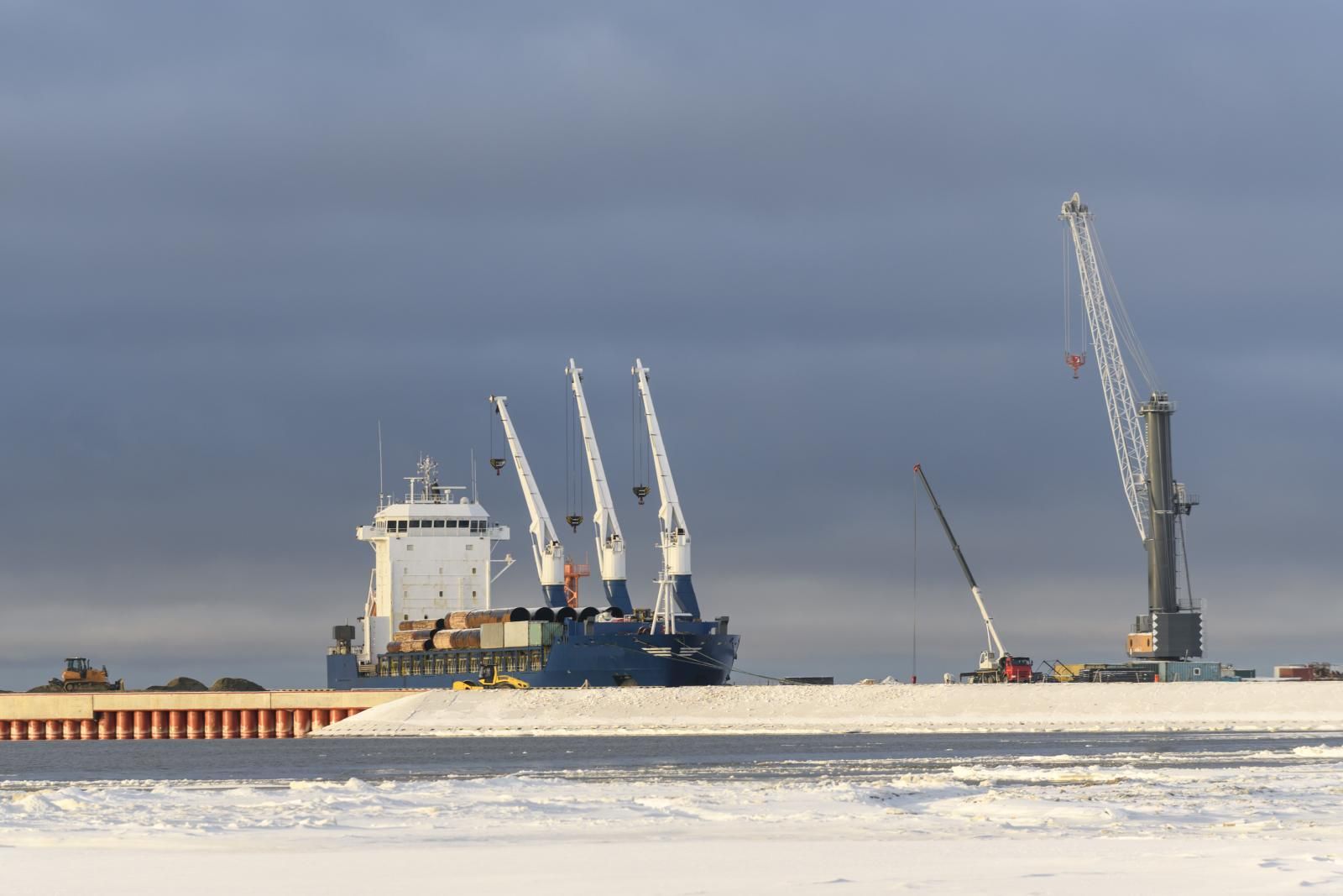 A cargo ship with deck cranes, loaded with steel pipes, timber and containers, moored at a snow-covered quay beside port cranes.