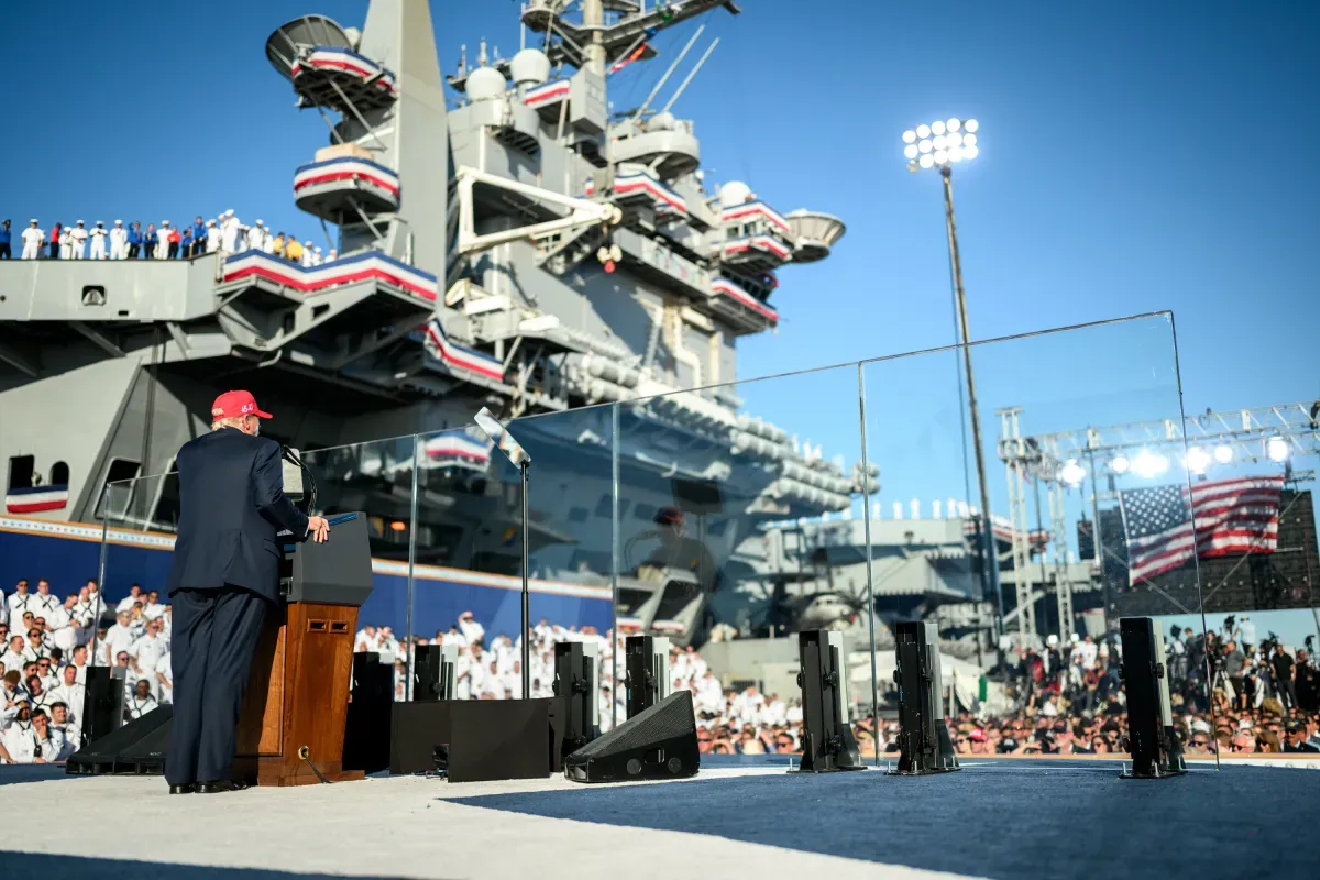 Donald Trump, de espaldas y con gorra roja, habla en un atril ante un portaaviones de la US Navy engalanado, con cientos de marineros formados en uniforme blanco de gala.