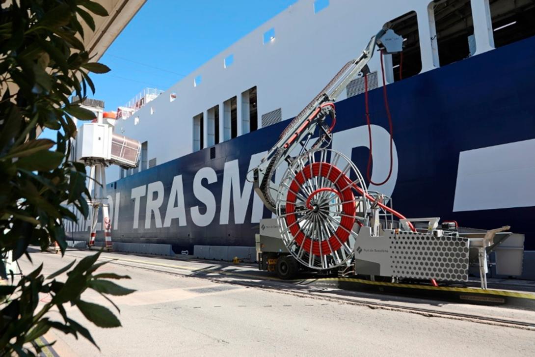 Una unidad móvil de electrificación OPS con un gran carrete de cable rojo, en el muelle, conecta con el casco de un gran ferri de Trasmed (Grimaldi) atracado bajo un cielo despejado.
