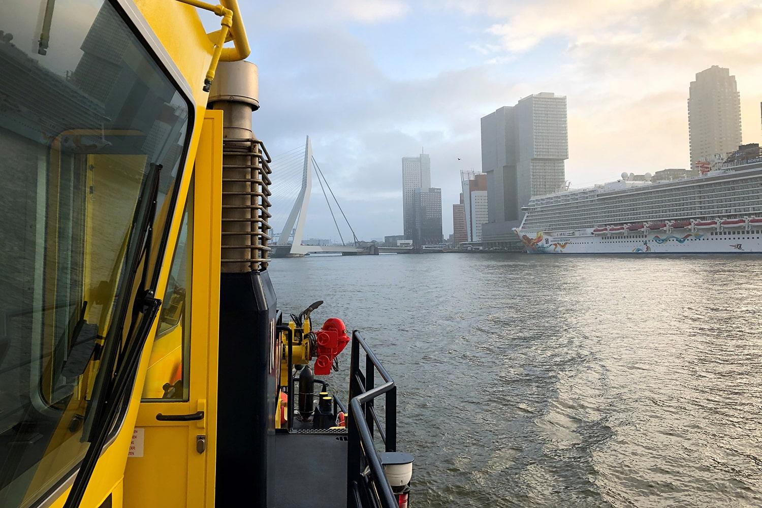 Desde la cubierta de una embarcación de vigilancia amarilla del puerto de Rotterdam se ve el skyline de la ciudad —el puente Erasmus y las torres de Kop van Zuid— y un gran crucero atracado junto a la orilla.