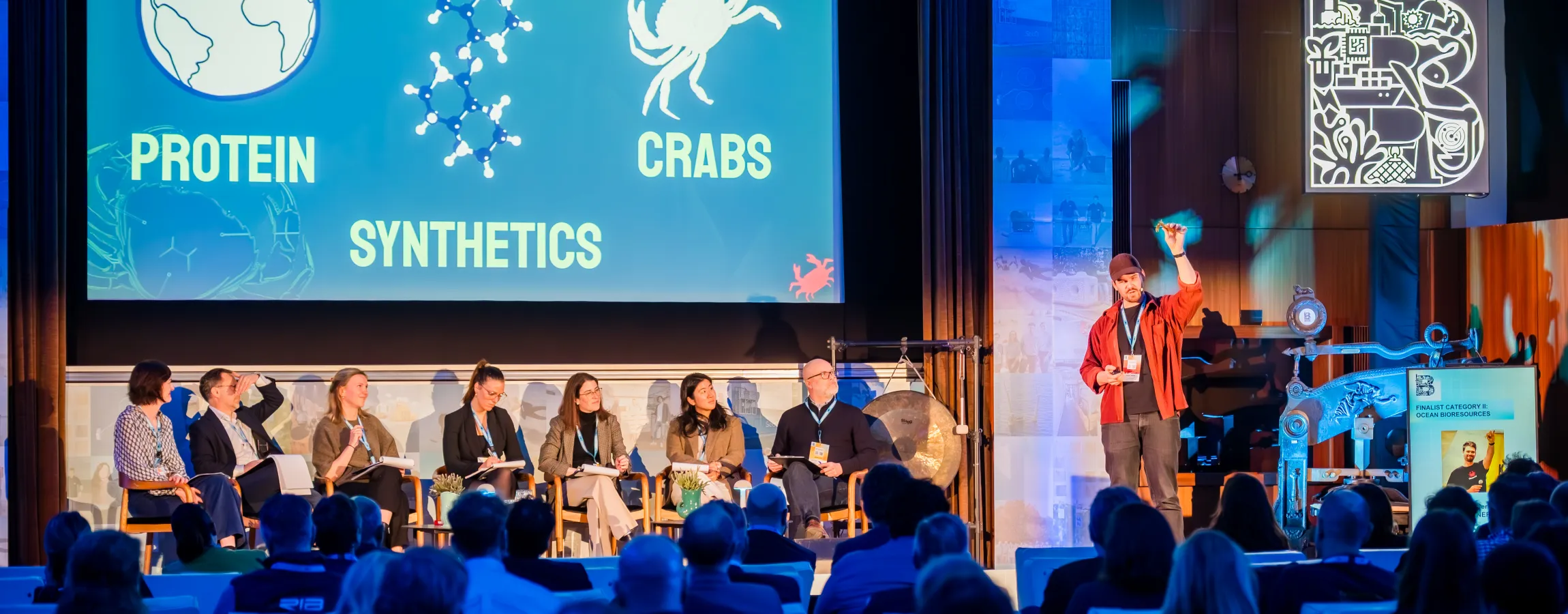 Sesión plenaria del BlueInvest Day 2026 en Bruselas con una pantalla que ilustra biotecnología azul y un panel de expertos sentado en el escenario.